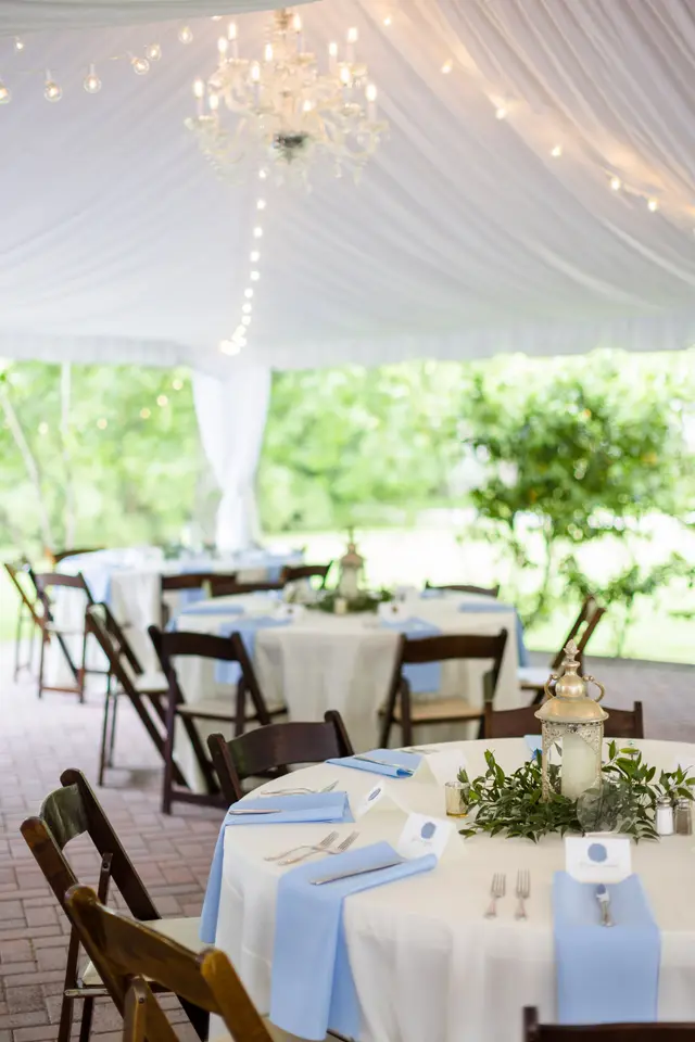 Round tables with blue napkins under a white tent and a chandelier
