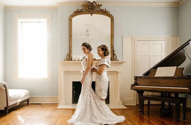 Bride being buttoned into her gown in the blue parlor by the grand piano
