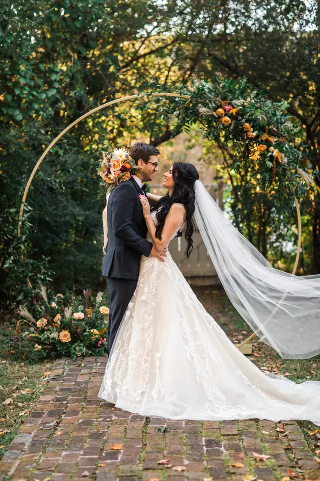 Couple kissing beneath a floral arch on the brick path, her veil trailing