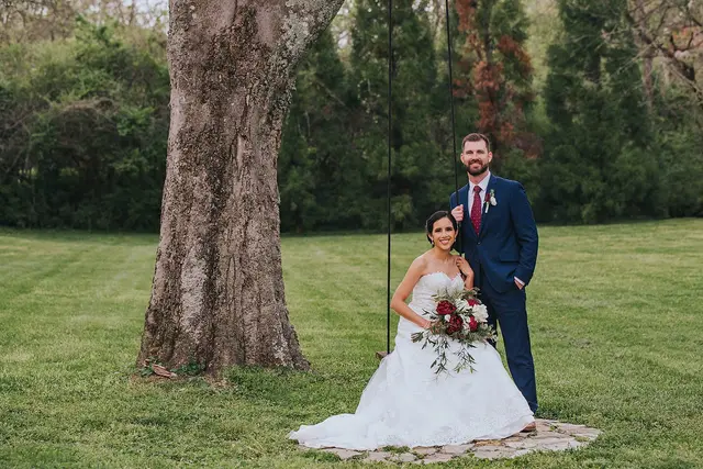 Couple under a big tree on the lawn, the bride holding a red bouquet
